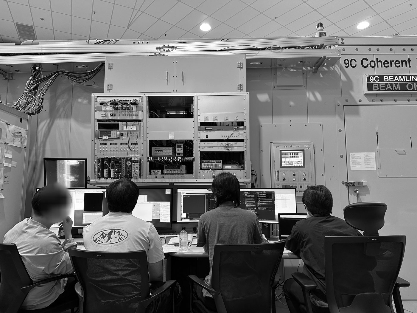 Four people seen from behind at the control desk of the 9C Coherent X-ray beamline, facing monitors under an electronics rack; a sign on the green hutch wall reads BEAM ON.